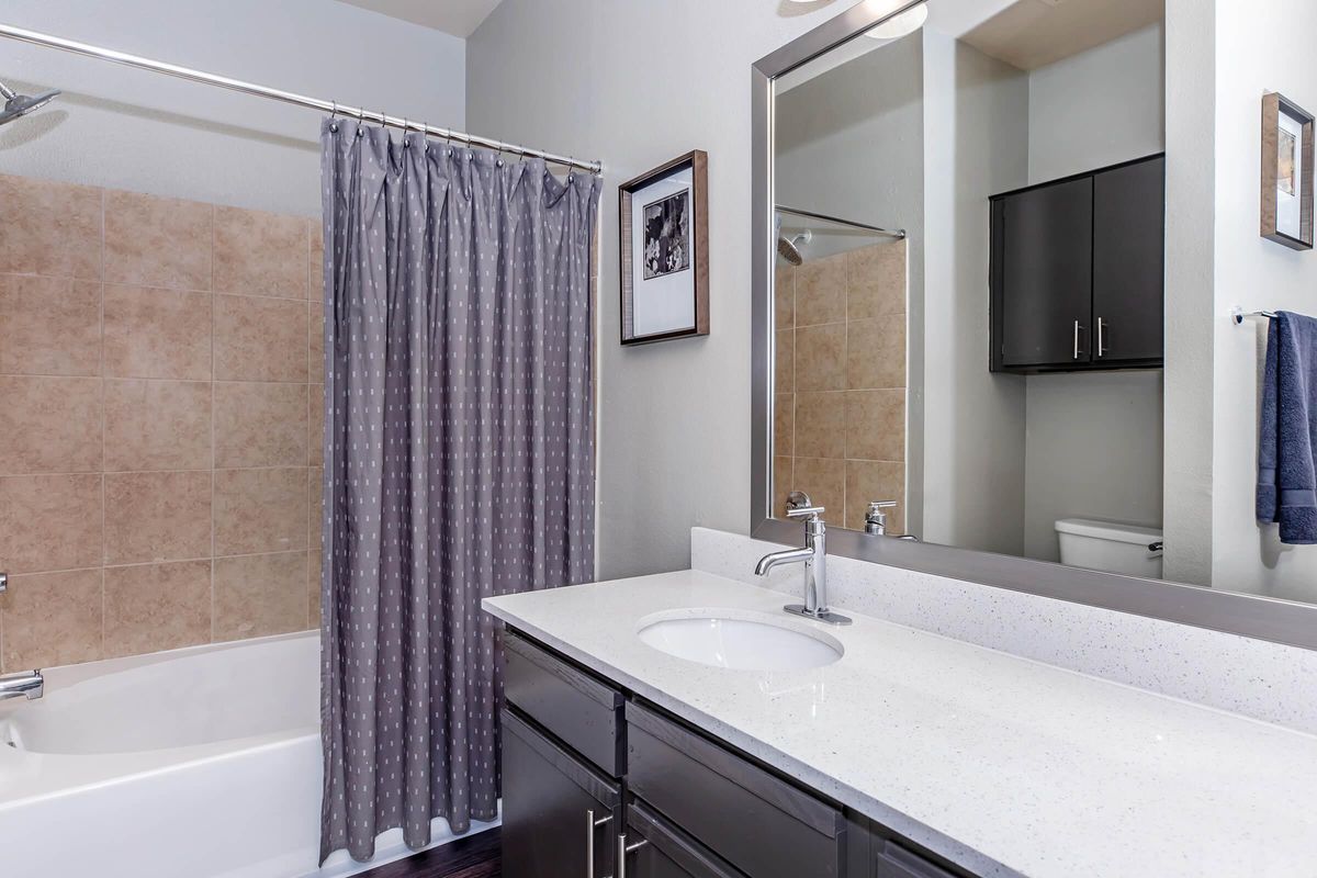 A modern bathroom featuring a bathtub with a shower, a gray polka dot shower curtain, and a white countertop with a sink. The walls are a light color, and there’s a large mirror above the sink, reflecting a storage cabinet and a towel hanging nearby.