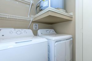 A laundry room featuring a white washing machine on the left and a white dryer on the right, both against a beige wall. Above the machines is a shelf with a small hot water heater. Wire shelving is mounted on the wall for storage. The space is well-lit and organized.