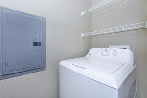 A white washing machine located in a laundry room, positioned next to a wall-mounted electrical panel. Above the machine are wire shelving units for storage, and the walls are painted in a neutral tone, creating a clean and organized space.