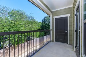 A balcony with a black door, surrounded by green trees and a clear blue sky. The railings are metallic and the floor is concrete. The walls are painted in a light green color, creating a serene outdoor space.