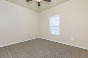 Empty room with light beige walls and carpeted floor. A small window with white blinds is located on one wall, and a ceiling fan hangs from the ceiling. The space is bright and minimalistic, with no furniture or decorations.