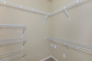 Empty closet with light beige walls and multiple white wire shelves arranged along the walls. The shelves are positioned at different heights, providing organized storage options. There are two electrical outlets visible on the left wall. The floor is carpeted in a neutral color.