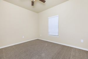A spacious room with light beige walls and beige carpet flooring. It features a ceiling fan, a single white window with closed blinds, and is largely empty with no furniture, creating a clean and open atmosphere.