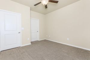 A vacant room with light beige walls and carpet. There is a ceiling fan with wooden blades, and two doors are visible: one leading to the closet and another possibly to a bathroom or hallway. The space is well-lit and minimalist, suitable for various purposes.