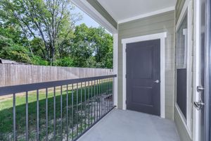View of a balcony area featuring a gray door, a railing, and a concrete floor. The background includes a grassy area bordered by a wooden fence and trees, suggesting a peaceful outdoor setting. Bright daylight illuminates the space, creating an inviting atmosphere.