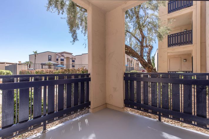 View from a shaded balcony with blue railings, showcasing a sunny courtyard area with greenery. In the background, a glimpse of another building can be seen, adding to the residential atmosphere. The scene is bright and inviting, with clear blue skies above.