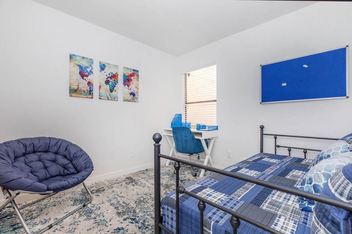 A cozy bedroom featuring a metal bed with blue and plaid bedding, a circular blue chair, and a desk with blue accessories. The walls are decorated with a colorful world map artwork and a blue notice board. Natural light enters through a window, illuminating the space.