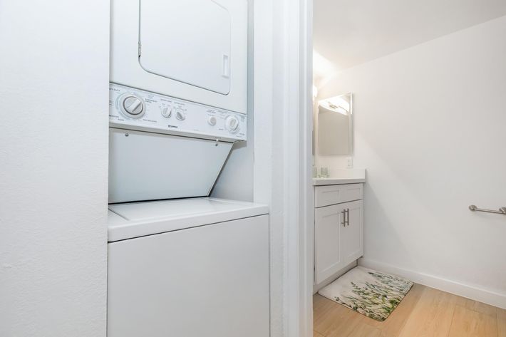 A compact laundry area featuring a stacked washer and dryer unit next to a modern bathroom. The bathroom includes a clean, white vanity with a mirror and is decorated with a green and white bath mat on a light wooden floor. The walls are painted in a bright, neutral color.