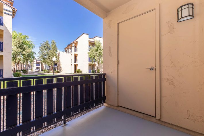 A view from a balcony featuring a closed door and a black railing. The background shows a landscaped courtyard with trees and nearby apartment buildings, under a clear blue sky. Sunlight illuminates the scene, creating a welcoming atmosphere.