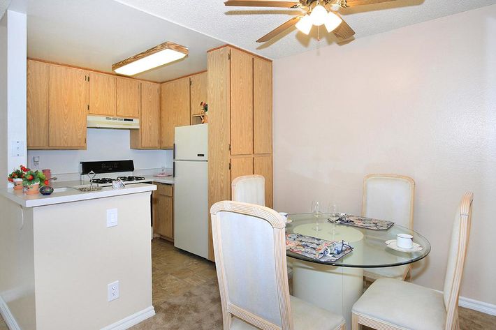 A cozy kitchen and dining area featuring light wooden cabinetry, a white refrigerator, and a stove. The round glass dining table is set for two, with floral placemats and cups. The space is bright and inviting with a ceiling fan and soft neutral wall colors.