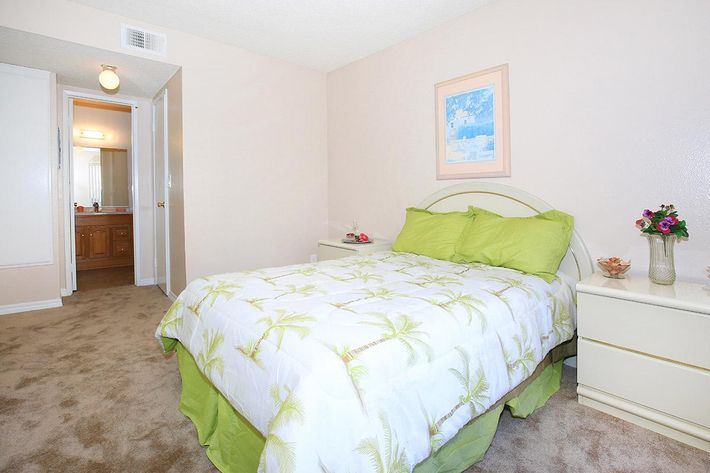 A cozy bedroom featuring a queen-sized bed with a light green and white floral quilt. The room has beige walls and carpet, and a small nightstand with a vase of flowers. A doorway leads to a bathroom, and a framed picture hangs above the bed, adding a touch of decor.