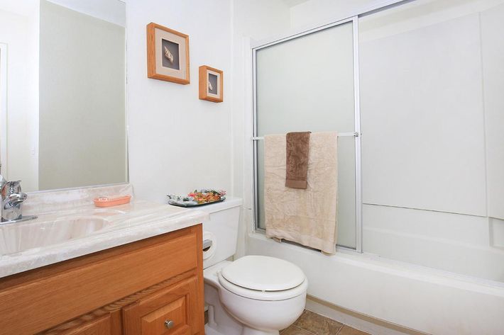 A clean, modern bathroom featuring a countertop with a sink, wooden cabinetry, and a mirror. A white toilet is visible, along with a shower area that has a sliding glass door. A towel hangs on a rack, and framed pictures adorn the wall, adding a touch of decor. The flooring is a light-colored tile.