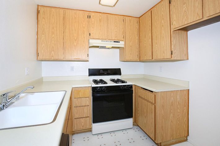 A small kitchen featuring wooden cabinets, a white countertop, a two-basin sink, and a black gas stove with an overhead range hood. The walls are painted light, and the flooring has a subtle pattern. The layout is compact, emphasizing a functional space.