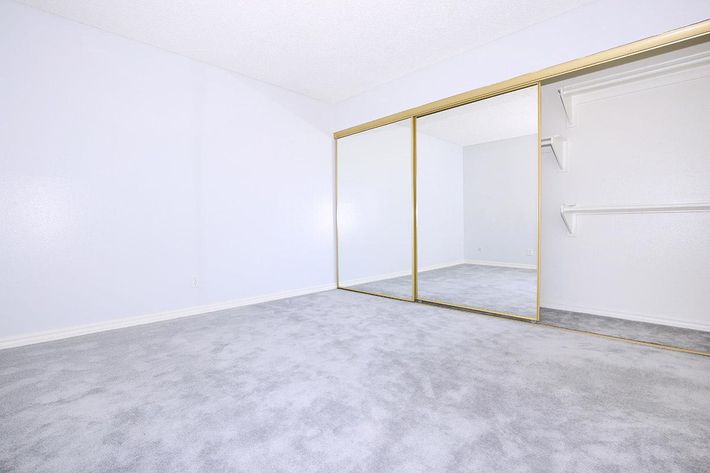 A bright, empty room with light gray carpet and pale blue walls. A large mirrored closet with gold trim occupies one wall. The space has a minimalistic feel, with no furniture or decorations present, creating a clean and open atmosphere.