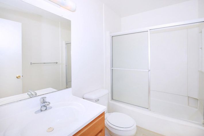 A clean and modern bathroom featuring a white color scheme, a glass shower enclosure, a white toilet, and a sink with a wooden vanity. The room has a large mirror above the sink and light colored flooring, creating a bright and spacious atmosphere.