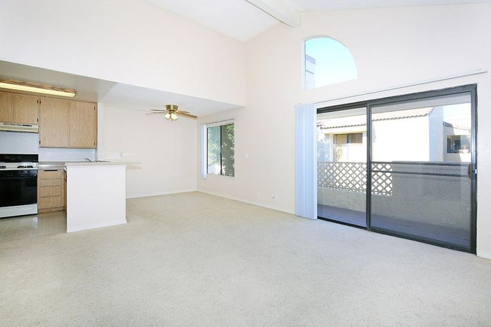 A spacious, well-lit living area with beige walls and carpet. The kitchen features wooden cabinets, a black stove, and a ceiling fan. A large window and sliding glass door lead to a balcony or outdoor space, enhancing natural light in the room. Ideal for a cozy home or apartment setting.