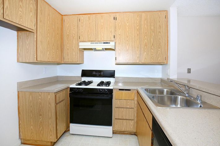 A kitchen featuring wooden cabinetry, a black gas stove with a hood, and a dual stainless steel sink. The countertops are light-colored laminate, and the overall design is functional and modern. The space is well-lit and organized, ideal for cooking and meal preparation.