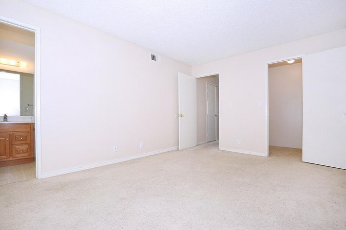 A spacious, empty room with beige walls and light-colored carpet. There are two doors visible: one leading to a bathroom with wooden cabinetry and a sink, and the other to a closet or another room. Natural light illuminates the space through an unseen window, creating a bright and airy atmosphere.