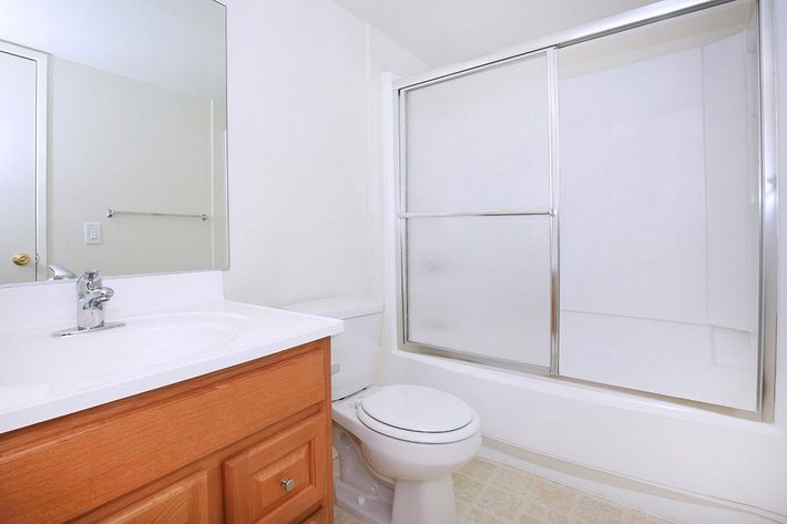 A clean, modern bathroom featuring a white sink with a chrome faucet, a toilet, and a glass-enclosed shower. The walls are light-colored, and there is a large mirror above the sink. The floor has a light, patterned tile, creating a bright and spacious atmosphere.