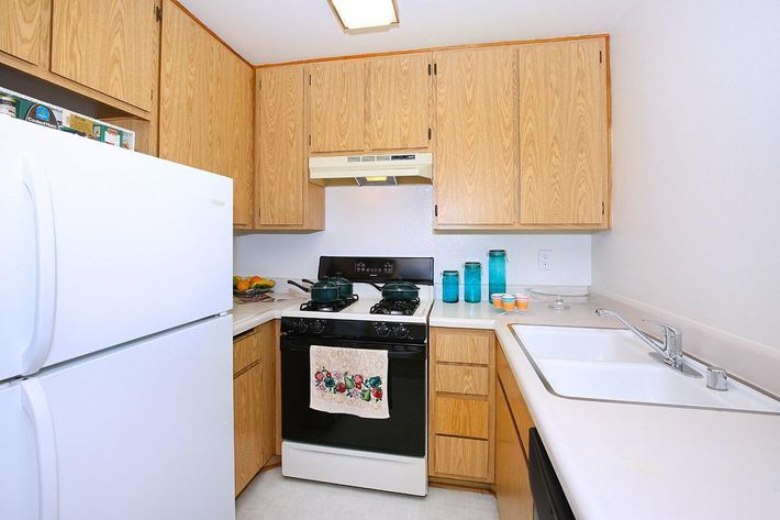 A small kitchen featuring wooden cabinets, a white refrigerator, a black stove with a hood, and a sink. The countertop is light-colored and there are decorative items, including colorful jars and a towel with a floral design. The overall space is bright and functional.