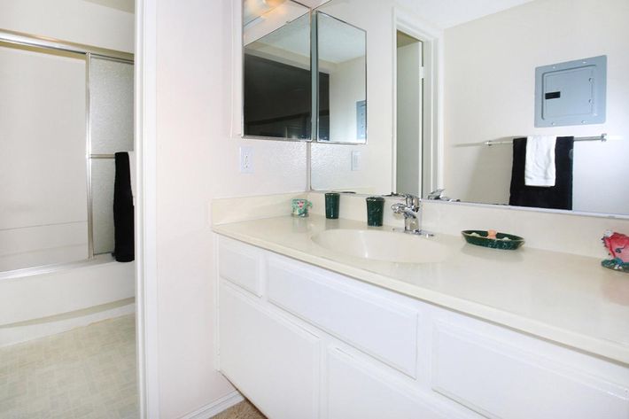 A clean bathroom featuring a light-colored countertop with a sink, a large mirror above, and decorative items. To the left, there's a shower area with a glass door, and a towel rack with a black towel. The walls are painted a soft color, creating a bright and spacious atmosphere.