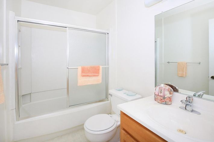 A bright, clean bathroom featuring a glass shower enclosure, a white toilet, and a sink with a wooden cabinet. Two peach-colored towels hang on a towel rack, and a decorative bag is placed beside the sink. The walls are painted white, contributing to a fresh and spacious atmosphere.