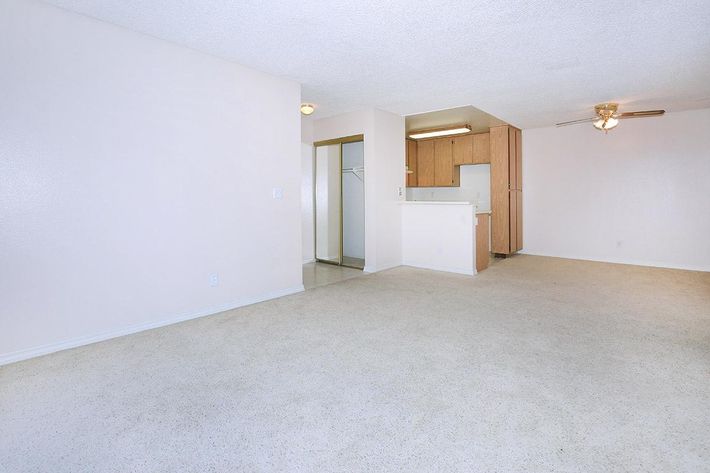 An empty living area with light-colored walls and carpet. The space features a ceiling fan and an open layout leading to a kitchen area with wooden cabinetry. The room is spacious and well-lit, with natural light coming from the adjacent area.