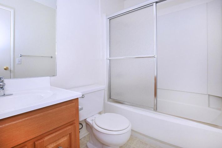 A clean and modern bathroom featuring a white countertop sink with wooden cabinetry, a toilet, and a shower-tub combination with a glass sliding door. The walls are light-colored, and there is a mirror above the sink. Natural light brightens the space.