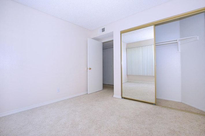 A spacious, light-colored room with beige carpeting. The room features a door leading to an adjoining space and a large closet with mirrored doors. There is a window with vertical blinds allowing natural light to enter, creating a bright, airy atmosphere. The walls are plain, enhancing the minimalist feel.