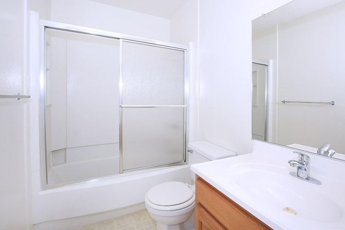 A clean and simple bathroom featuring a shower with glass sliding doors, a white toilet, and a sink with a wooden cabinet underneath. The walls are painted white, and there is a mirror above the sink reflecting the bright, minimalistic design. The flooring is light-colored vinyl.