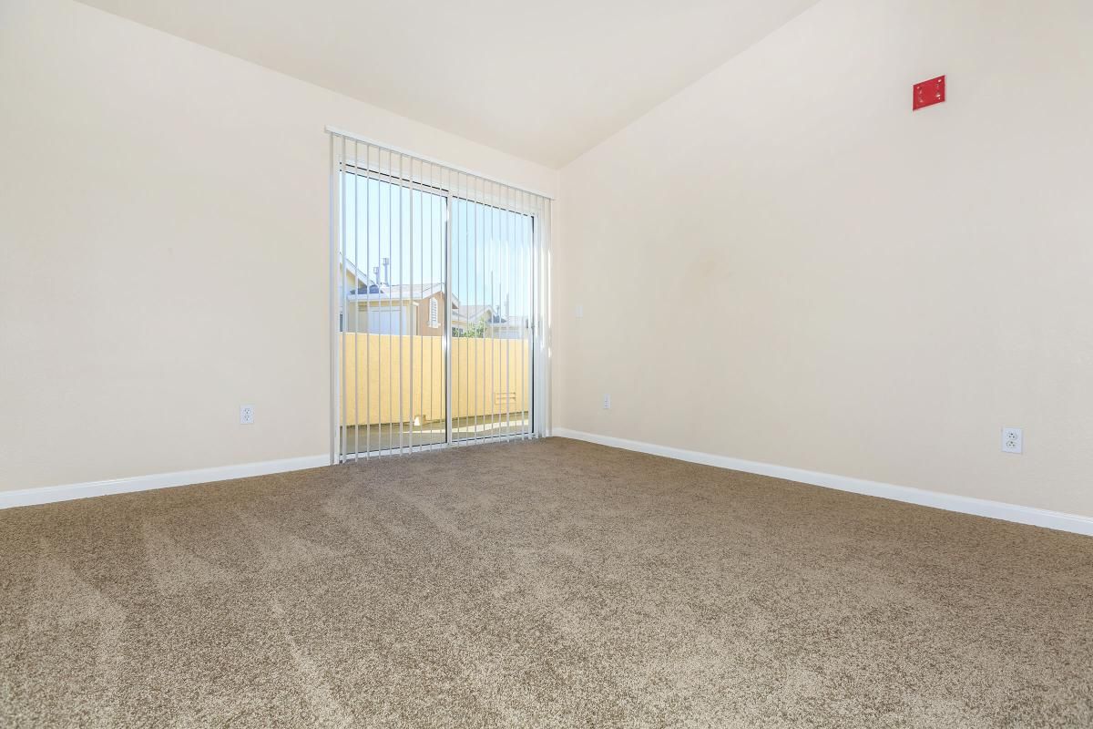 A spacious, empty room with light beige walls and a large window covered by vertical blinds. The floor is a soft, light brown carpet, and there's a red fire alarm on one wall. The view through the window shows a yellow balcony railing outside. The overall ambiance is bright and airy.