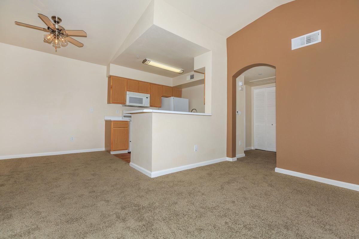An interior view of a compact, unfurnished living space featuring light-colored walls and a brown accent wall. The kitchen, with wooden cabinets and a white refrigerator, is partially visible. The room has a ceiling fan and beige carpet, with an archway leading to a hallway with a closet. Natural light fills the space.