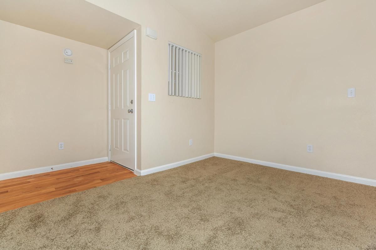 A vacant room featuring beige walls and carpeted flooring. There's a white door on the left leading to the entrance, and a small window with bars above. The space is bright and uncluttered, emphasizing the neutral tones of the interior.