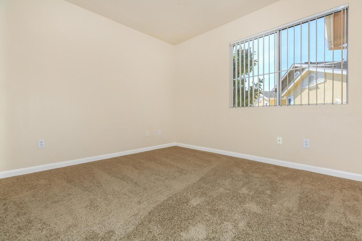 A vacant room with beige walls and carpet. The room features a window with vertical blinds allowing natural light to enter. The space is empty, suggesting it's ready for new furniture or decoration. There's one visible wall outlet and no other furnishings or decorations present.
