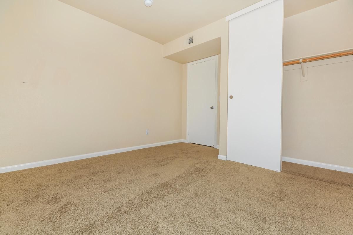 A vacant room with beige walls and plush, light brown carpet. There is a white closet with sliding doors on the left and a closed door leading to another space on the right. The ceiling is plain with a light fixture, and the overall atmosphere is bright and neutral.