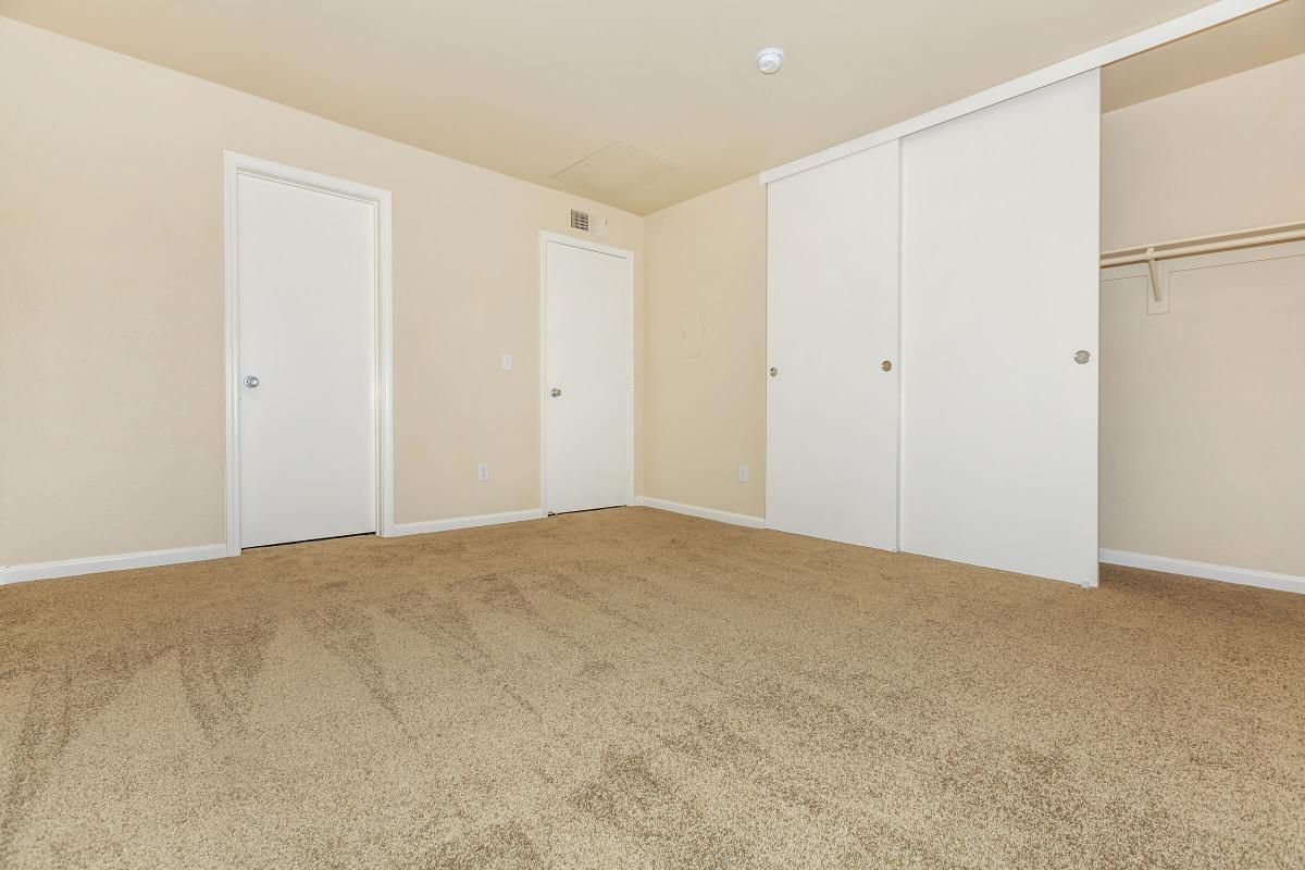 A spacious, empty room with beige walls and carpet. Two closed doors are visible on the left side, and a large closet with sliding white doors is on the right. The room has a neutral, light appearance, and there are no furnishings or decorations present.