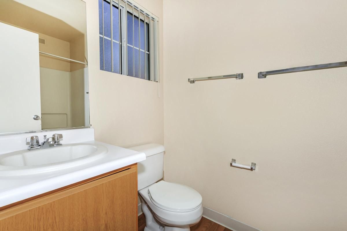 A clean bathroom featuring a white toilet, a sink with two faucets, and a mirror above the sink. The walls are a soft beige color, and there's natural light coming in from a window. The floor is wooden, and there are two towel bars mounted on the wall.