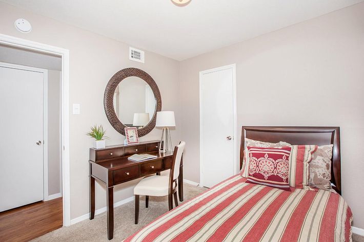 A cozy bedroom featuring a single bed with striped bedding, a wooden desk with a chair, a round mirror above the desk, and a small plant on the desk. The walls are painted in a light color, enhancing the room's brightness. A closed white door is visible in the background.