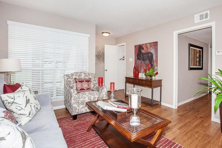 A cozy living room featuring a light gray sofa with patterned cushions, a wooden coffee table with decorative items, and two red candles. There is a small console table against the wall, adorned with a vase of flowers, and art pieces hang on the walls. Natural light filters through white wooden blinds.