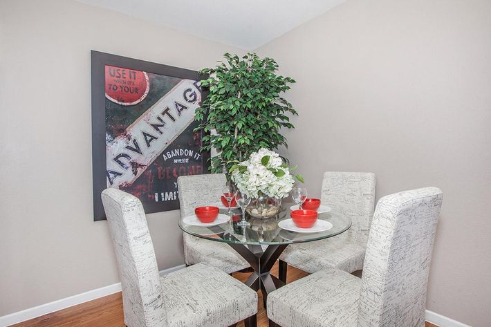 A cozy dining area featuring a round glass table with four light-colored, upholstered chairs. The table is set with two red bowls and a centerpiece of white flowers in a glass bowl, complemented by a decorative piece underneath. A large framed artwork hangs on the wall, and a potted plant adds greenery to the space.
