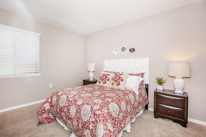 A cozy bedroom featuring a queen-sized bed with a red and cream patterned duvet, two bedside tables with lamps, and a small potted plant. The walls are painted a soft beige, and a window with white blinds allows natural light to brighten the space. Decorative mirrors are arranged above the headboard.