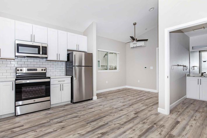 A modern kitchen featuring stainless steel appliances including a microwave, oven, and refrigerator. There are white cabinets with a tile backsplash and light wood-like flooring. The open layout connects to a well-lit living area with a ceiling fan and a view of a bathroom area in the background.