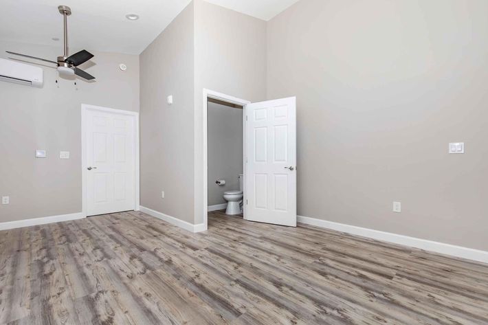Empty room featuring light gray walls and a wooden floor, with a ceiling fan, an air conditioning unit, and two closed doors. One door leads to a small bathroom, visible at the back. The space is well-lit, creating an inviting atmosphere.