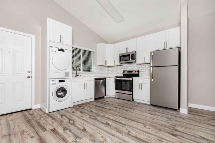 A modern kitchen featuring white cabinets, stainless steel appliances including a refrigerator, stove, microwave, and a washer-dryer unit. The room has a light-colored wall and a wood-grain floor, with a window providing natural light. The entrance door is visible on the left.