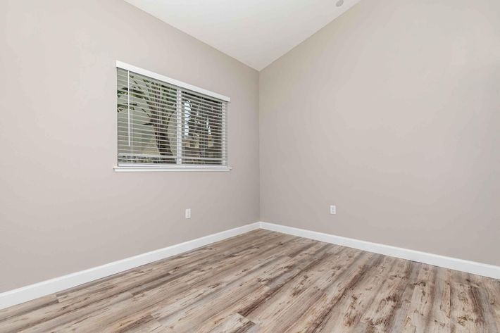 Empty room with light gray walls and wooden flooring. A single window with white blinds allows natural light to enter. The room features a high ceiling, creating an open and airy feel. There are no furnishings, making it suitable for various uses such as an office or a guest room.
