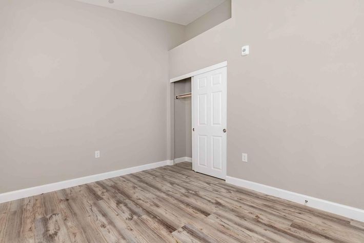 A spacious room with light gray walls and a high ceiling. The floor is covered with light-colored wooden planks. There is a white closet with double doors on the left side, and the overall atmosphere is bright and inviting, with no furniture present in the space.
