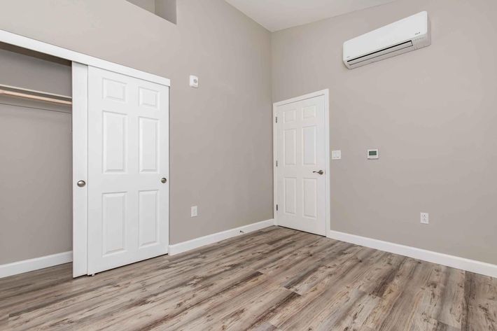 Empty room with light gray walls, a wooden floor, and an air conditioning unit mounted on the wall. There are two doors: one is a closet with sliding white doors, and the other is a standard white door. The space is well-lit and uncluttered, creating a clean and modern appearance.