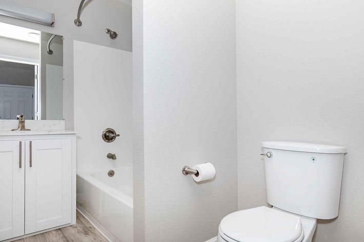 A modern bathroom featuring a clean design. The space includes a white toilet, a bathtub with a showerhead, a sink with a faucet, a mirror above the sink, and light-colored walls and flooring, creating a bright and open atmosphere. A roll of toilet paper is visible next to the toilet.