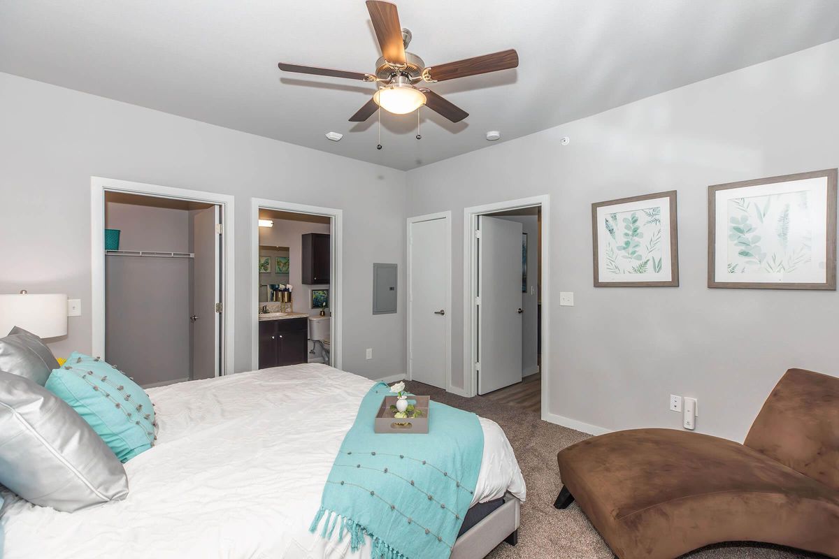 A modern bedroom featuring a queen-sized bed with gray and teal accents, a ceiling fan, and two doors leading to adjacent rooms. A cozy brown chaise lounge is positioned beside the bed, and artwork of green plants adorns the walls. The room has neutral-colored walls and carpet, creating a warm atmosphere.