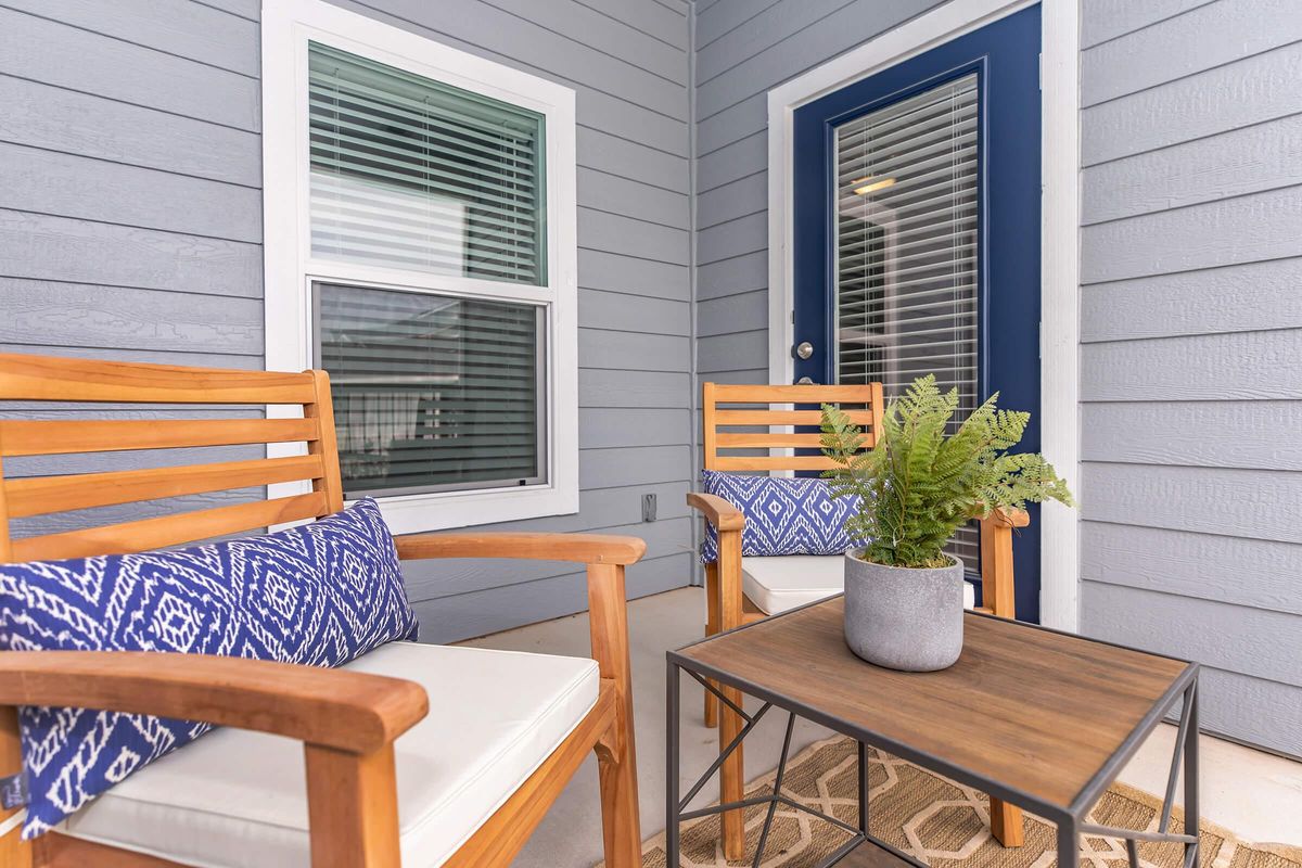 Two wooden chairs with white cushions and blue patterned pillows are positioned around a small brown coffee table. A potted fern sits on the table. The scene is set on a porch with light gray walls and a blue door, framed by large windows with blinds.